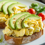 A mouthwatering plate featuring fluffy scrambled eggs with creamy avocado slices, served on golden toasted sourdough. The eggs have a light, soft texture, glistening slightly from the butter. The avocado, perfectly ripe, adds a vibrant green contrast. A sprinkle of black pepper and a few chilli flakes give the dish a hint of heat. Bright cherry tomatoes and a sprig of fresh coriander are delicately placed on the side, adding colour and freshness to the presentation. A drizzle of lime juice glistens on the avocado, making the whole dish look both hearty and inviting.